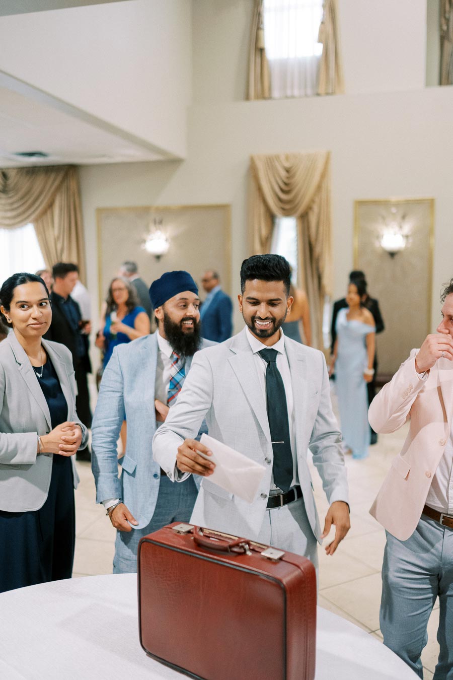 A group of well-dressed people at an indoor event, one man smiling while placing a letter into a red briefcase. They are in a room with elegant decor, including draped curtains and wall sconces, with other guests in the background.