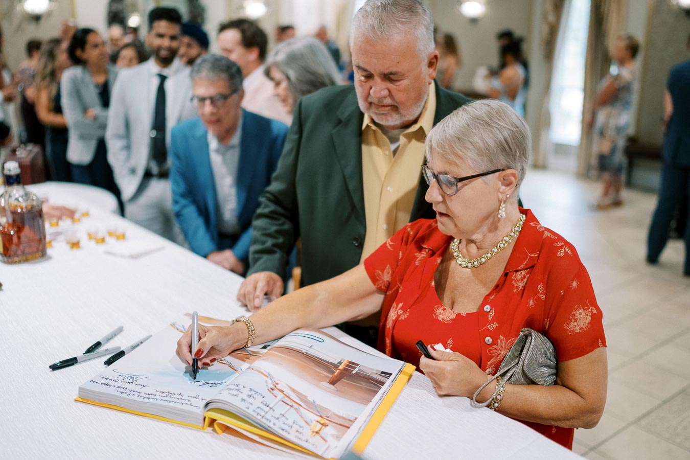 Elderly woman in red dress signing a guest book at a formal event, surrounded by attendees in a well-lit venue.