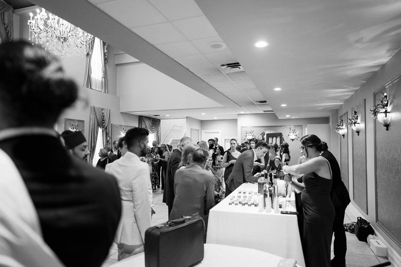 Black and white image of a lively indoor event with a group of well-dressed people gathered around a bar area, featuring elegant chandeliers and decorative wall sconces.