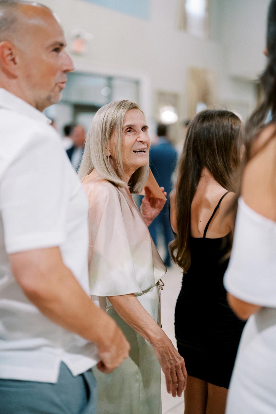 A group of people dressed in formal attire engaged in conversation at a social event. The setting appears to be a well-lit indoor venue.