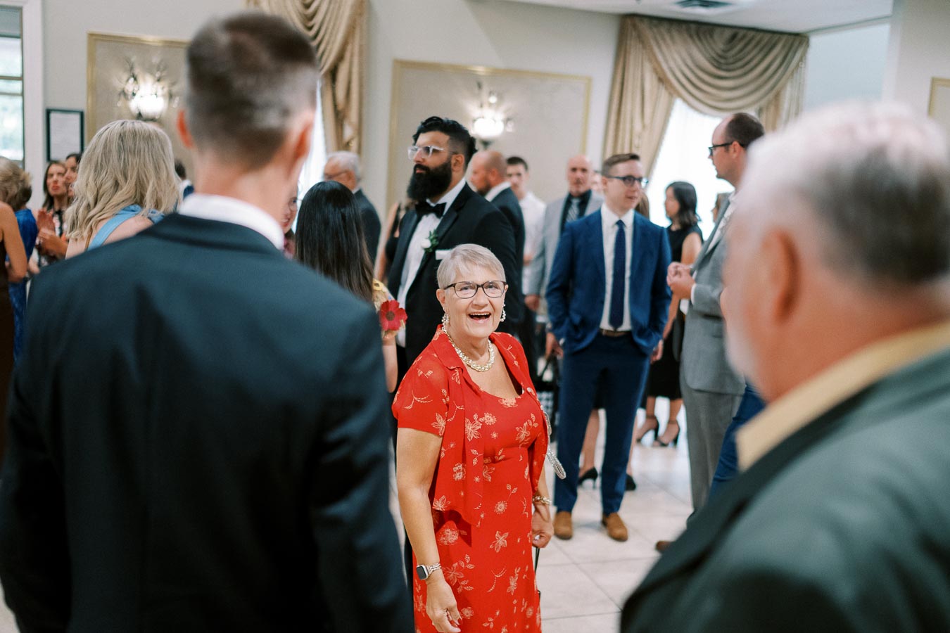 A lively social gathering with a woman in a red floral dress smiling among a crowd of elegantly dressed people at an indoor event.