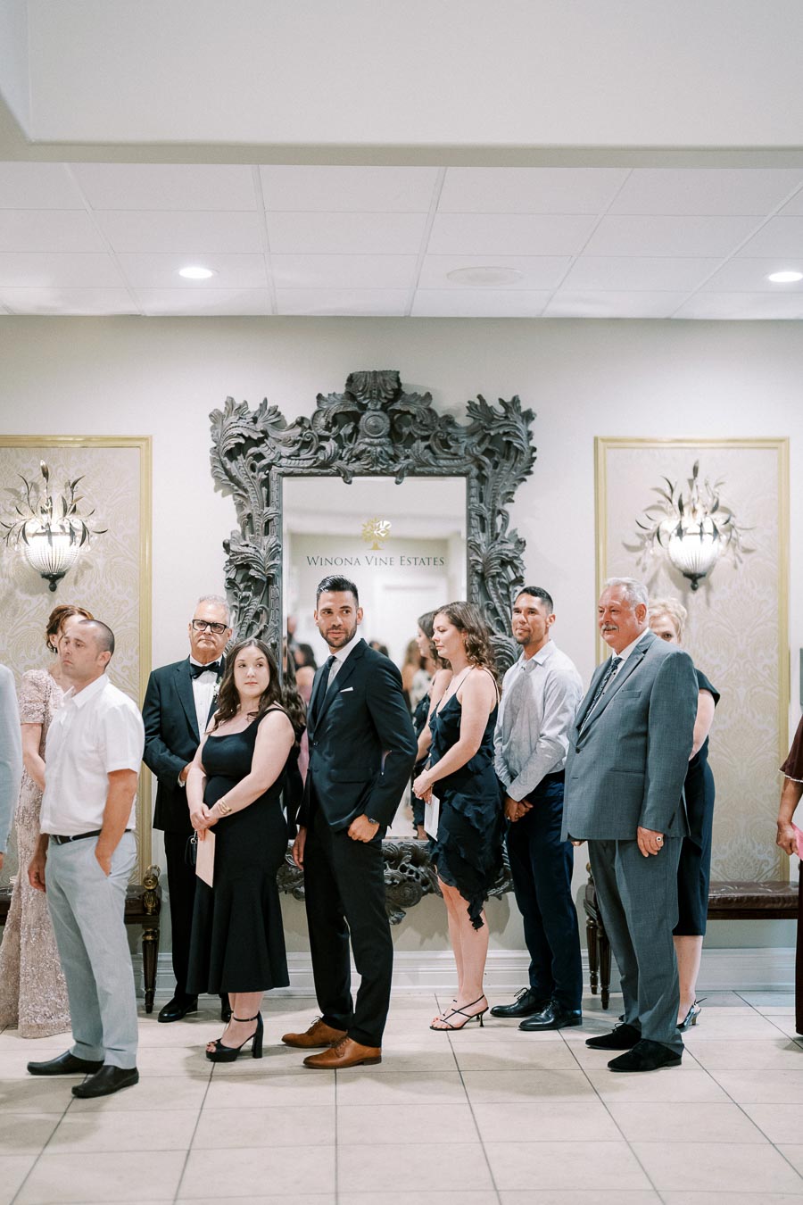 A group of well-dressed people standing in line indoors, with a decorative mirror labeled Winona Vine Estates on the wall.