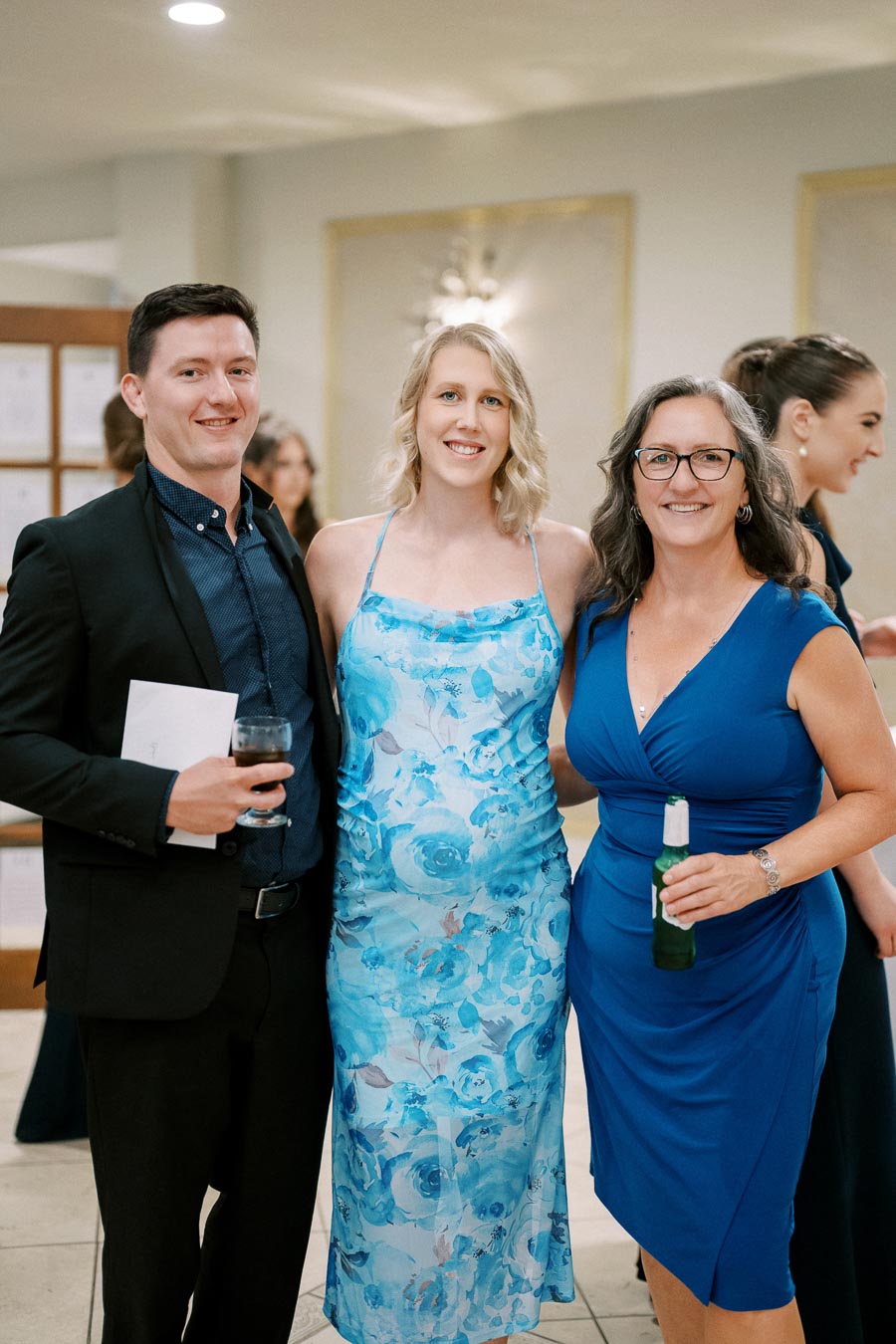 Three people at a social event, dressed formally, smiling and holding drinks.