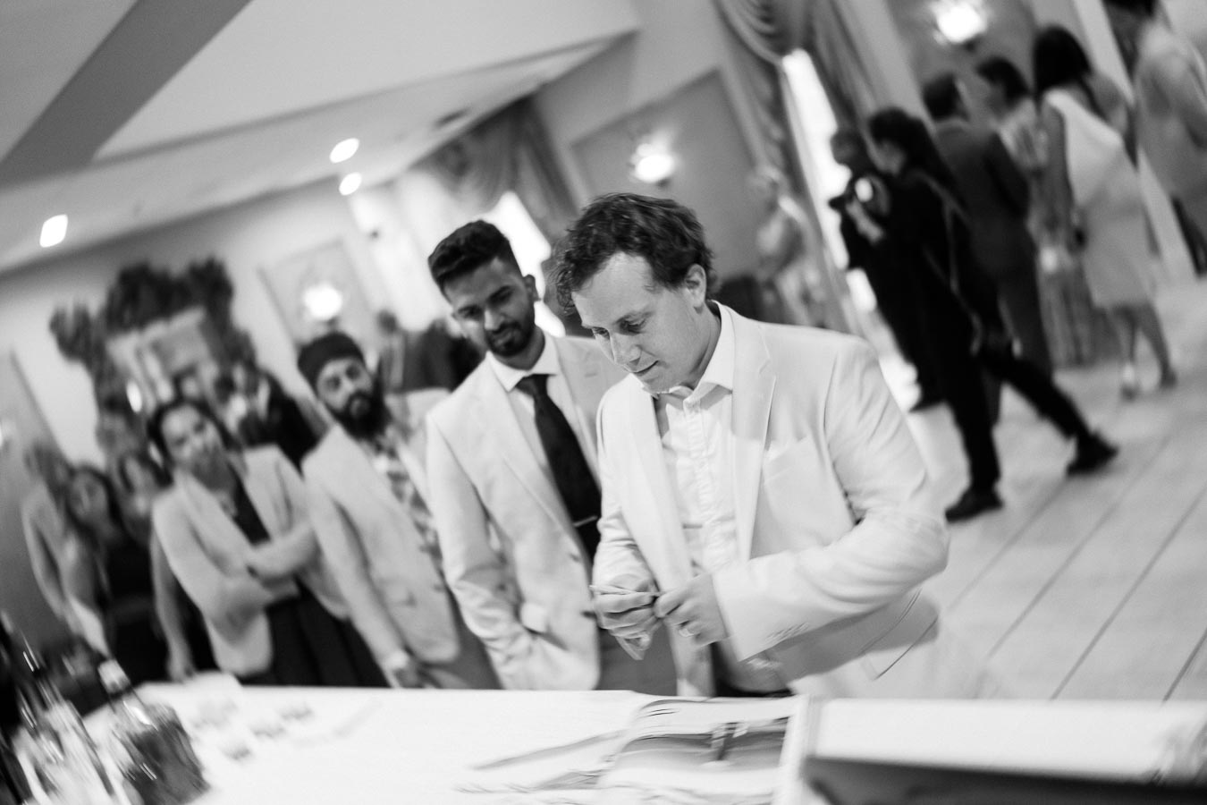 Black and white photo of a group of people in formal attire gathered at a reception desk during an event or conference, with blurred guests mingling in the background.
