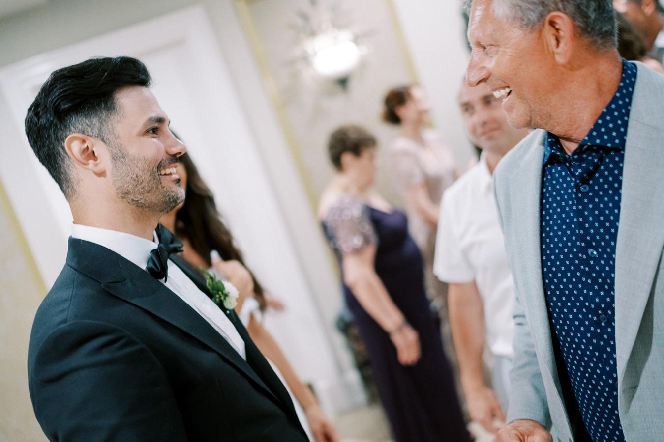 Smiling man in a black tuxedo conversing with another man in a gray suit at a formal event, surrounded by elegantly dressed guests.