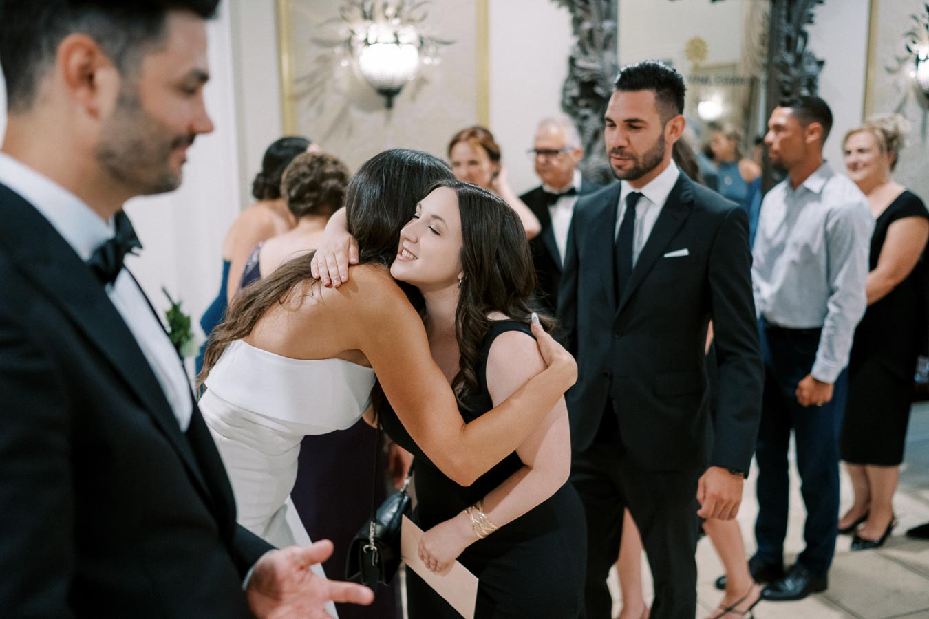 A wedding reception scene with guests in formal attire, two women hugging warmly, surrounded by elegantly dressed attendees, all in a beautifully decorated venue.