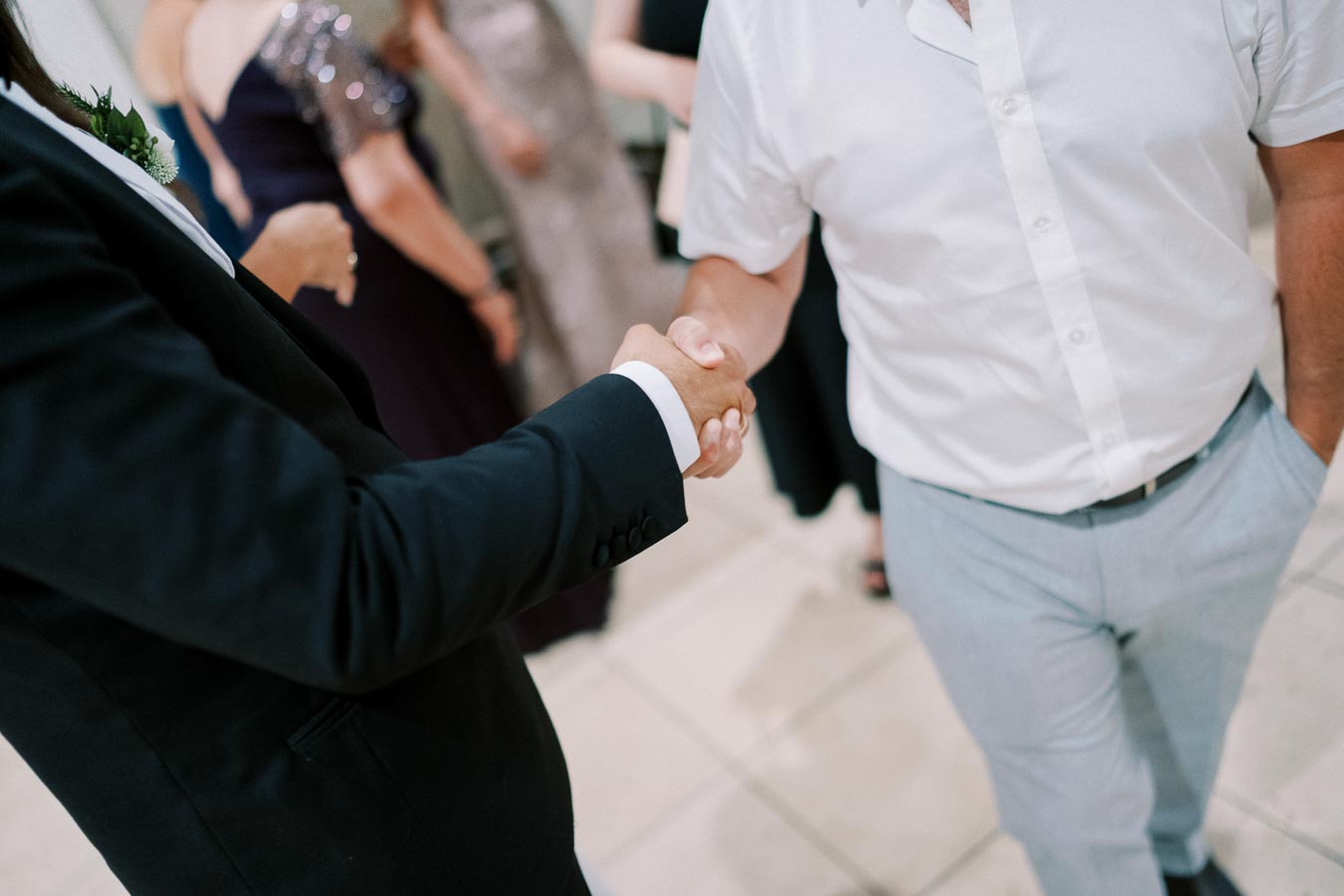 Two people in formal attire shaking hands at an event, signifying greeting or agreement, with other attendees blurred in the background.