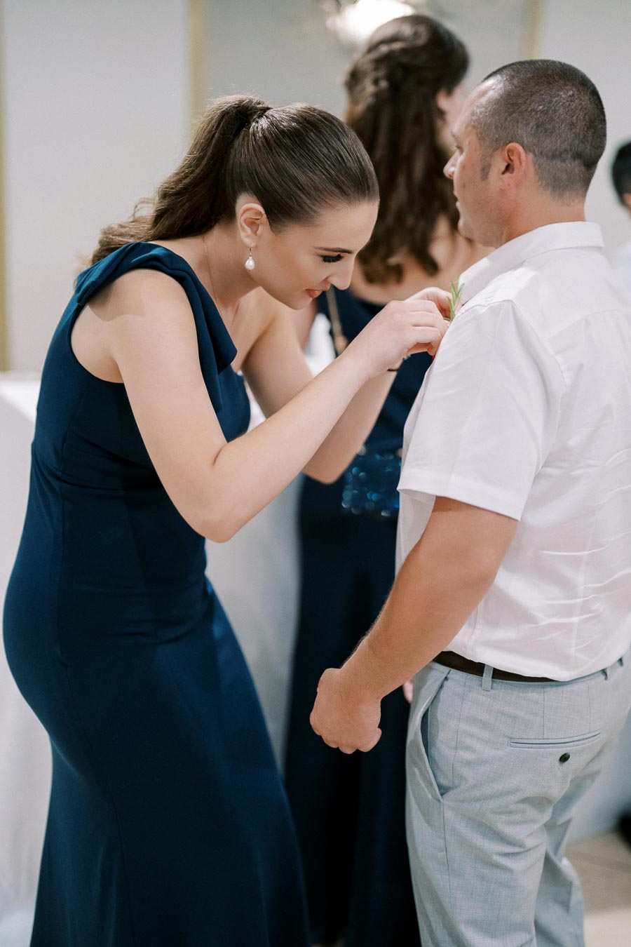 Woman in a navy blue dress adjusting a boutonniere on a man's white shirt at an event.