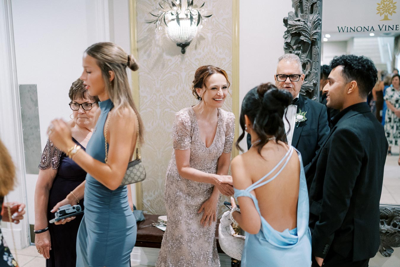 A group of elegantly dressed people socialize at a formal event at Winona Vine, with a woman in a lace dress greeting others.