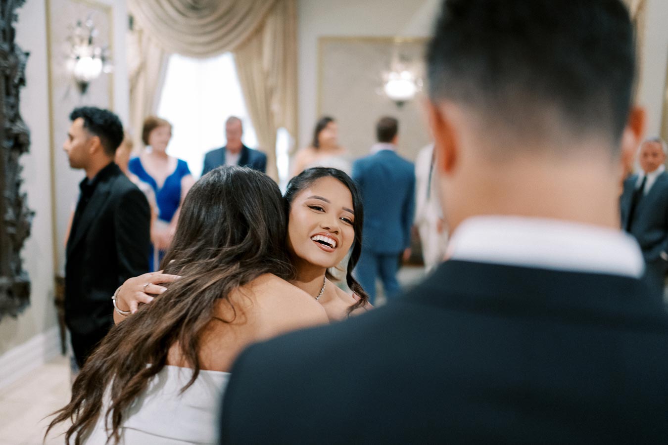 A joyful woman hugs a friend in a formal gathering, with elegantly dressed guests chatting in a lavishly decorated room in the background.