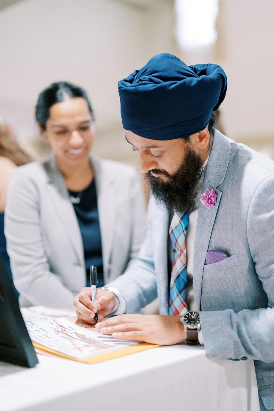 A man in a blue turban and light gray suit is signing a document, while a woman in a business outfit looks on, creating a professional setting.