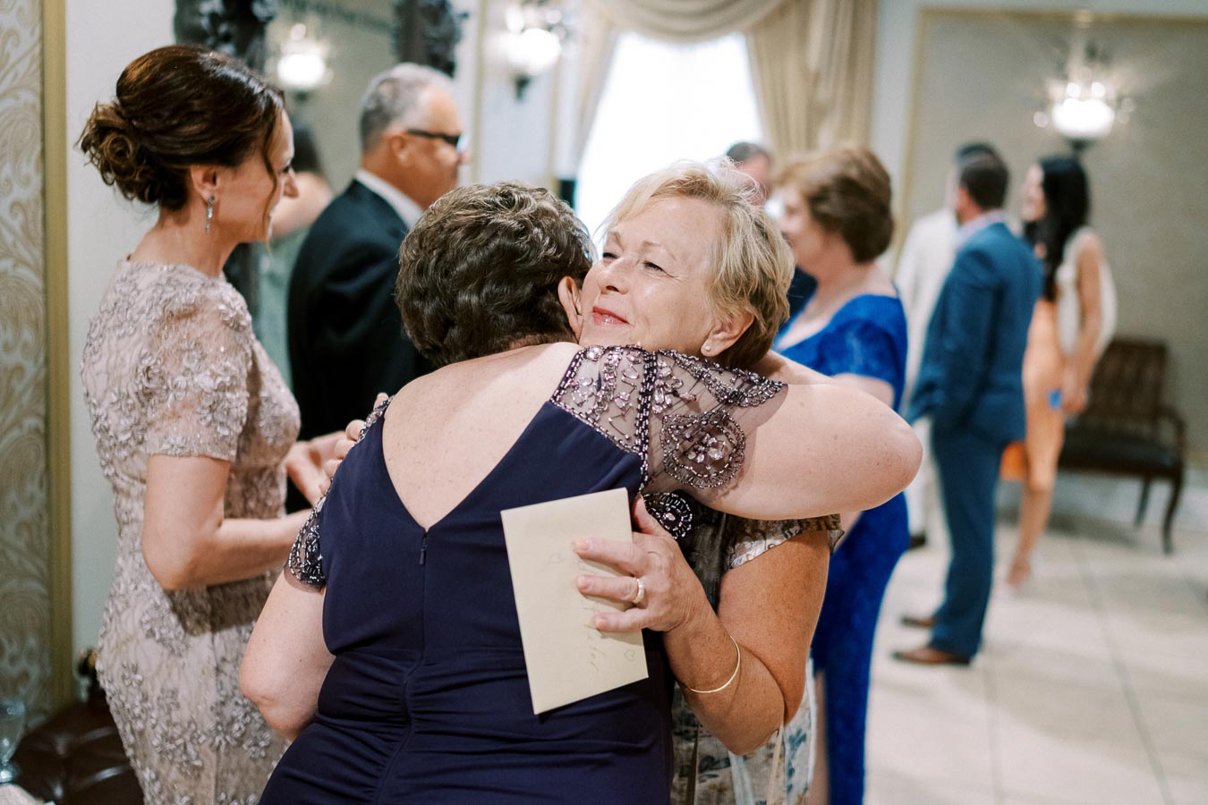 Elegant women embracing at a formal gathering, sharing a joyful moment, with other guests in the background, creating a warm and festive atmosphere.