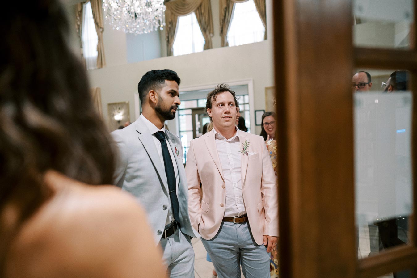 Two men dressed in light-colored suits, standing in a brightly lit, elegant room with chandeliers and draped curtains, engaged in a conversation during a formal event.