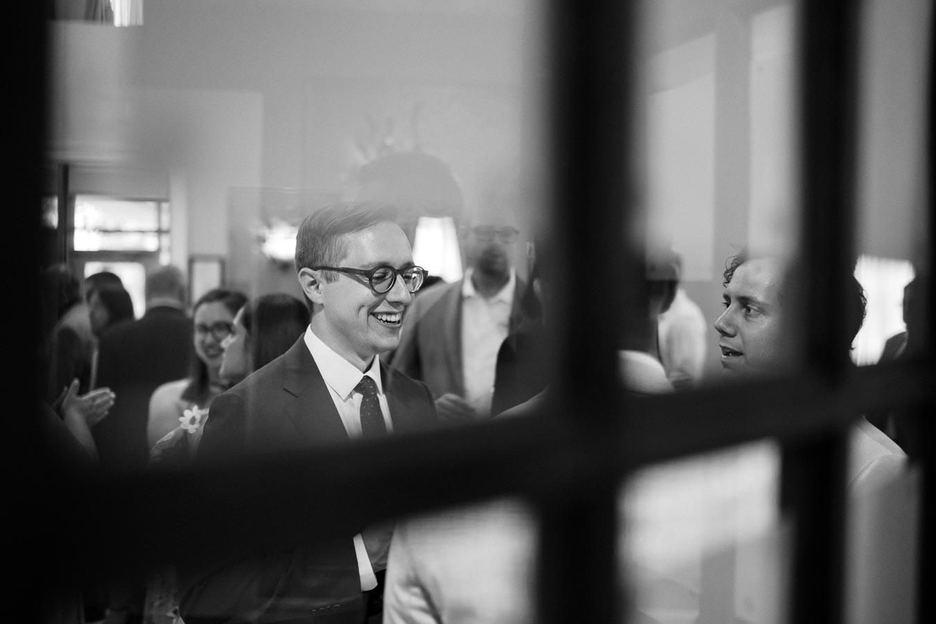 Black and white photo of people socializing at an indoor gathering, with a man in glasses smiling in the foreground, viewed through a window for an artistic perspective.