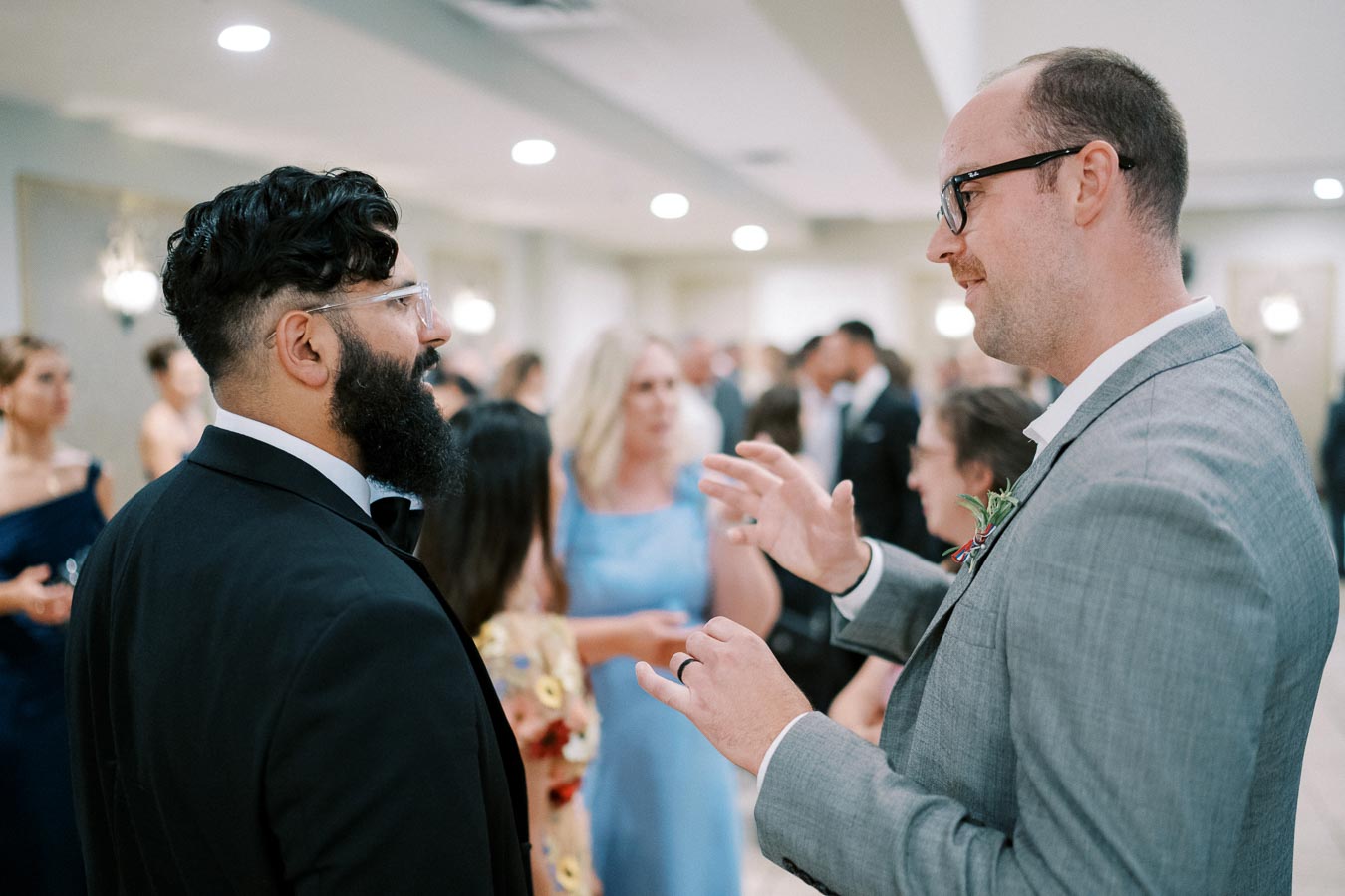 Two men in formal attire having a conversation at an elegant indoor event, surrounded by other well-dressed guests.
