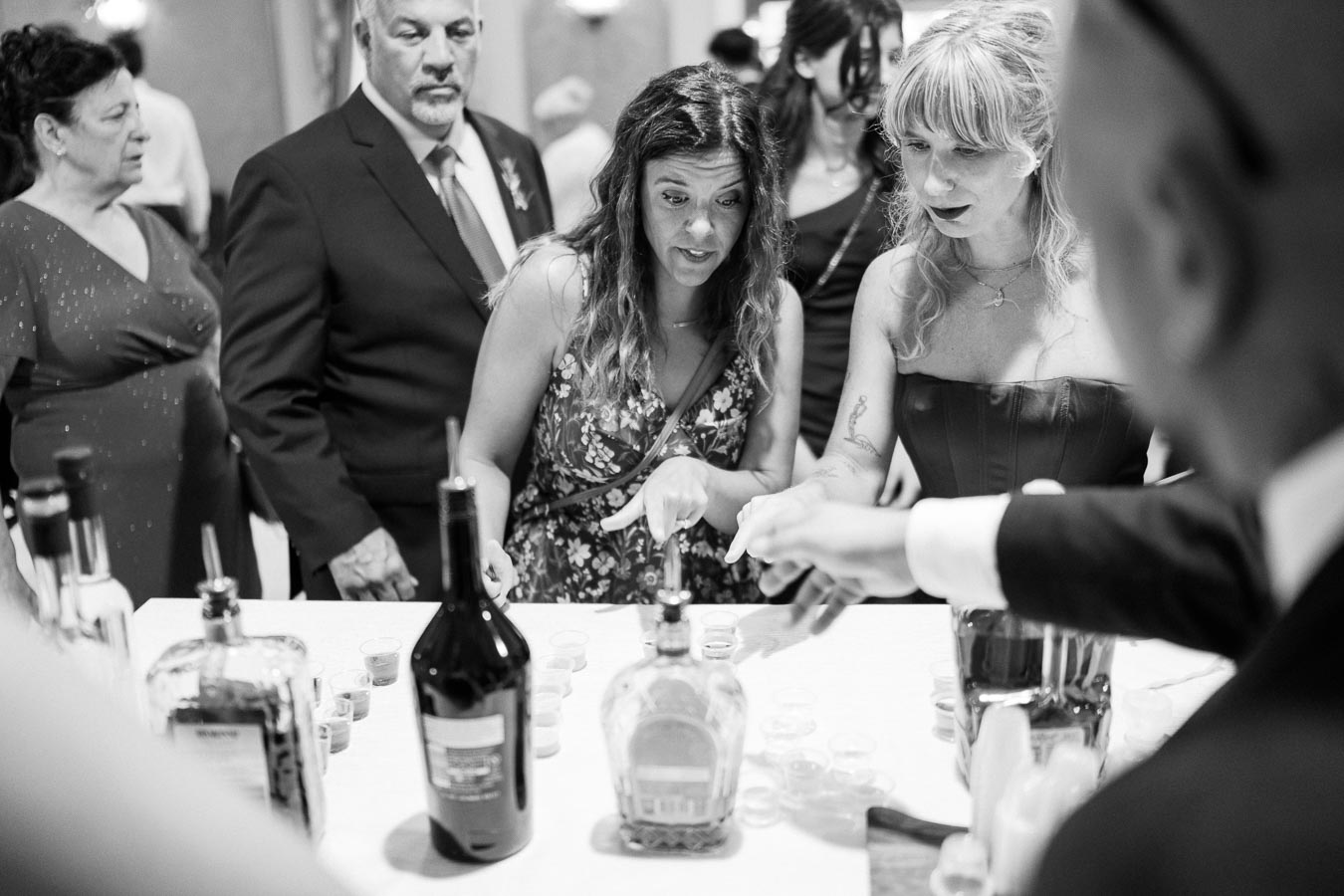 Black and white photo of a group of people gathered around a table with various bottles and shot glasses, engaged in conversation at a social event, possibly a wedding reception.