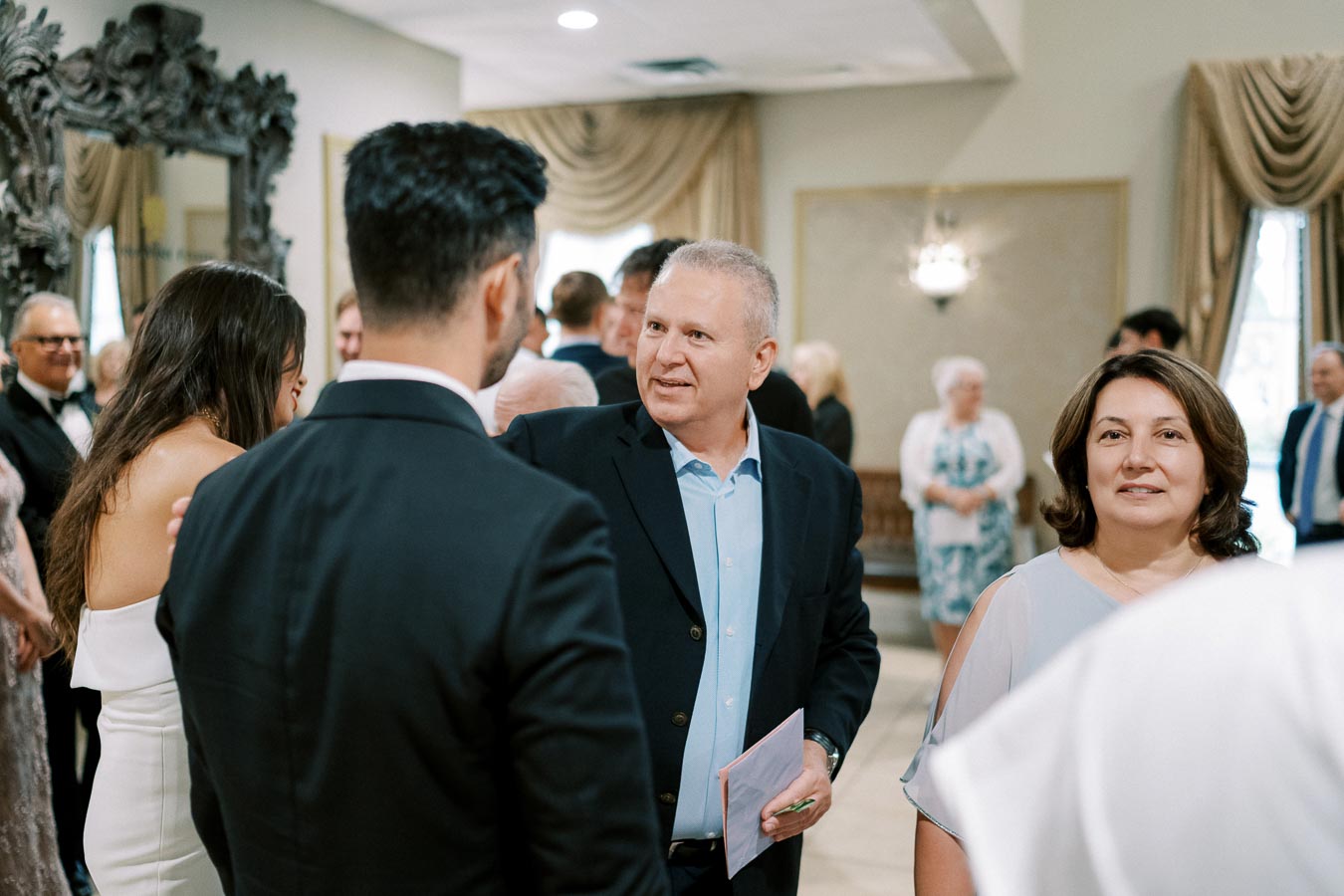 Group of people in formal attire engaged in conversation at an elegant indoor event, with a decorative mirror and curtains in the background.