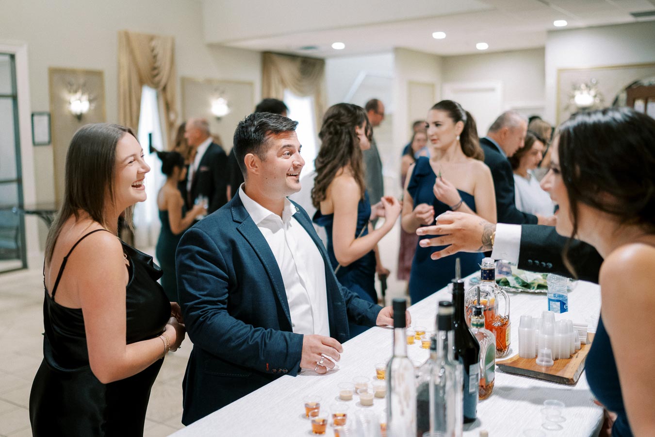 A lively social gathering at a well-decorated venue, featuring elegantly dressed individuals engaging in conversation near a bar with assorted bottles and cups.