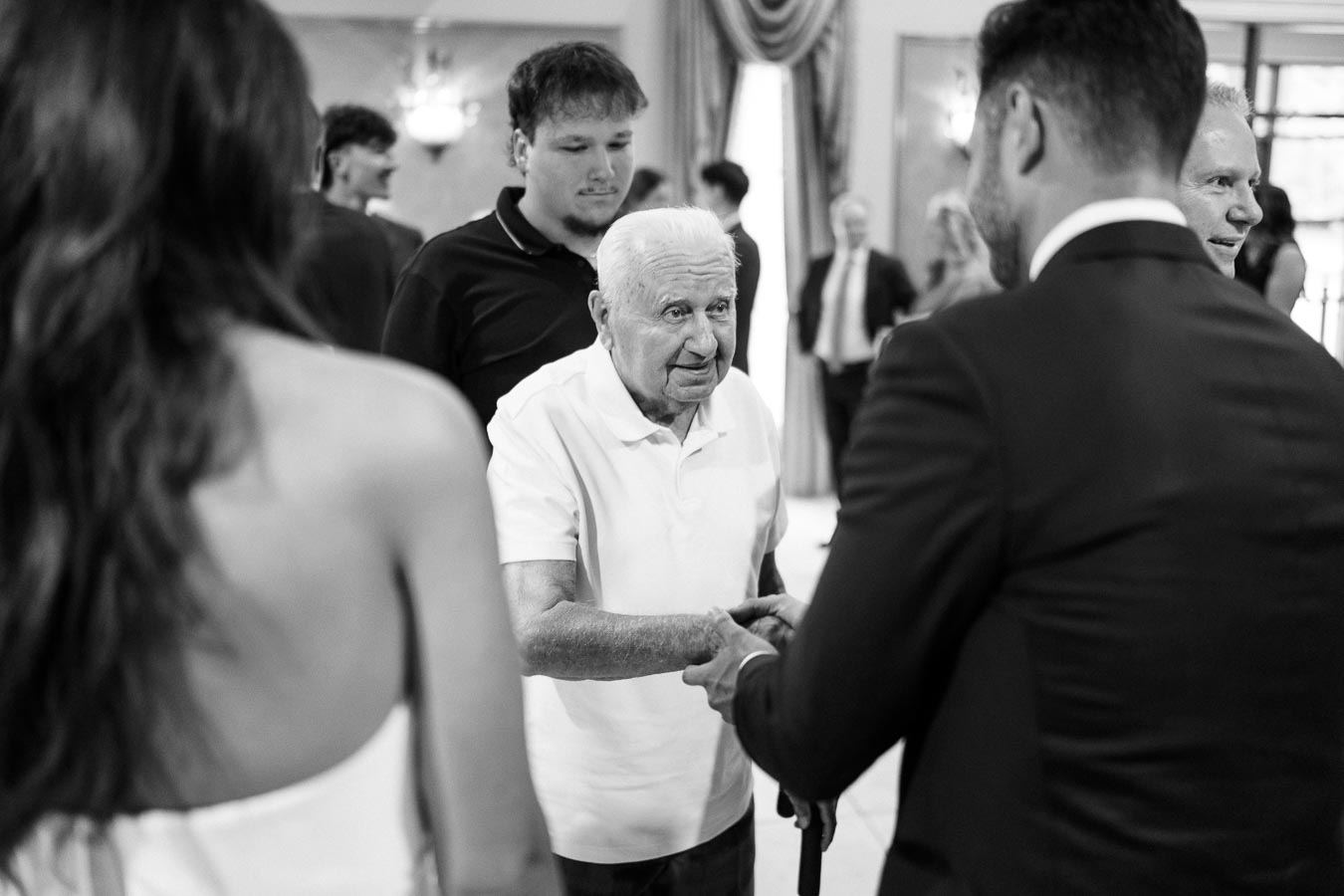 Black and white image of an elderly man in a white shirt greeting people at an indoor social gathering, shaking hands with a man in a suit while others stand nearby, in a warmly lit room with elegant curtains.