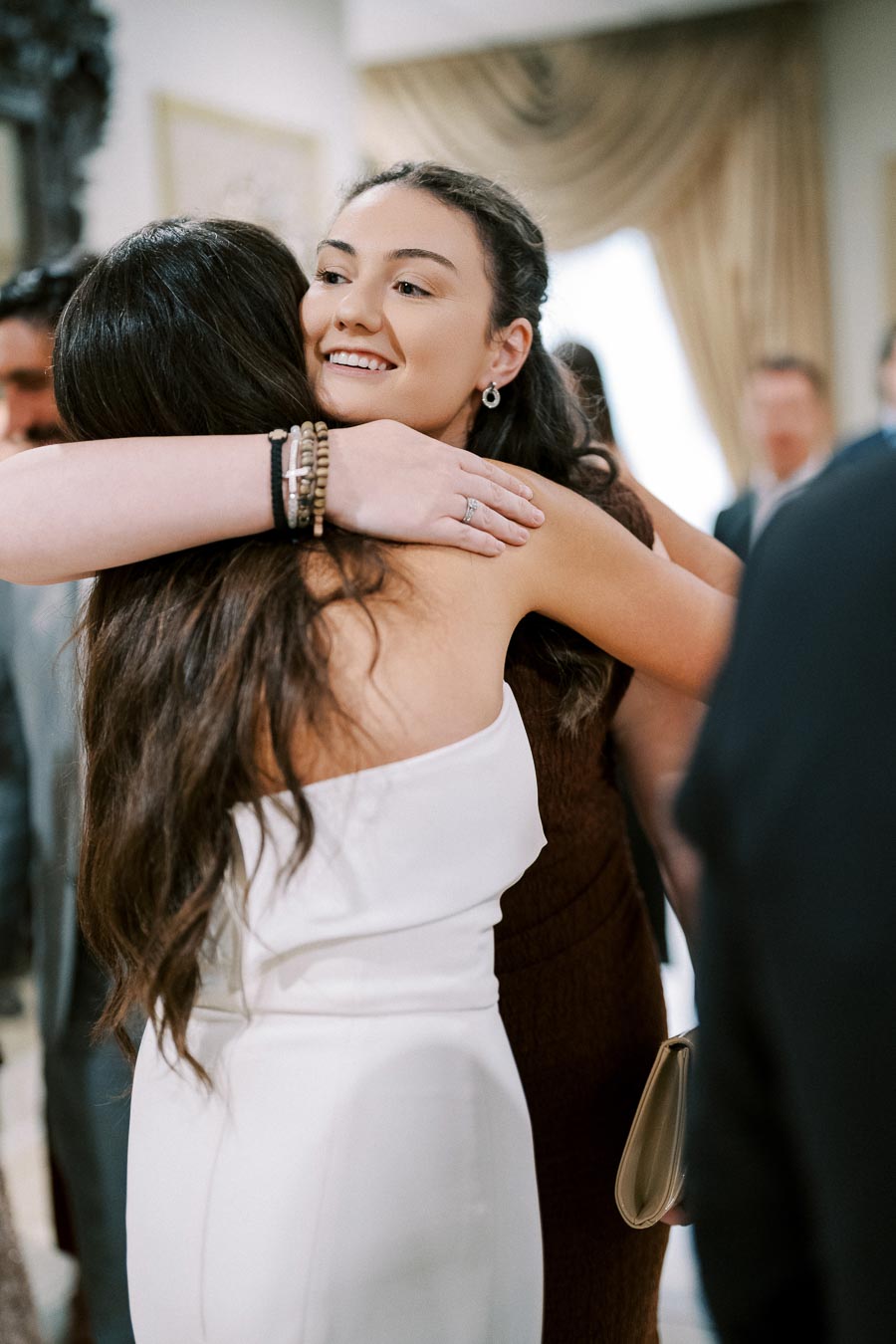 Two women embracing warmly at an indoor event, with one in a white dress and the other in a brown outfit. Background shows blurred attendees and elegant décor.