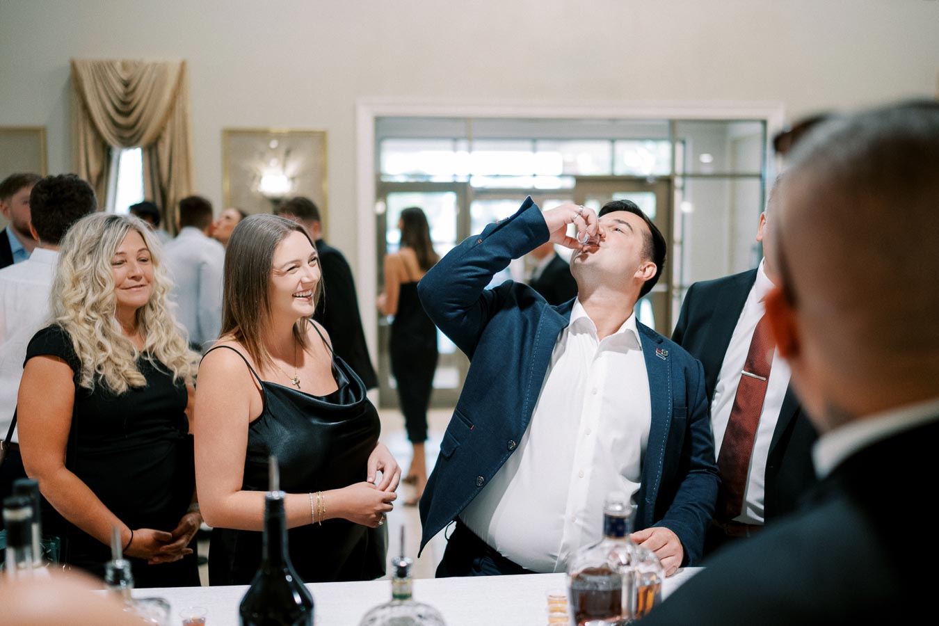 A group of well-dressed people enjoying a celebration, with a man taking a shot at a lively social gathering.