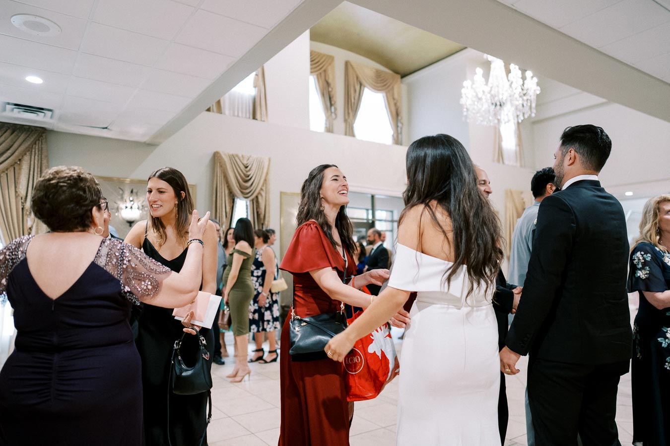 A group of elegantly dressed people socializing at an indoor event, featuring high ceilings, chandeliers, and draped curtains, creating a sophisticated and festive atmosphere.