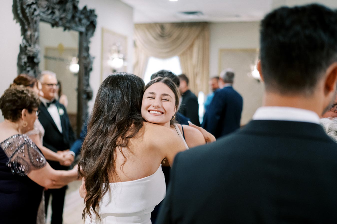 A joyful woman in a formal dress embracing another guest at a wedding reception, surrounded by elegantly dressed attendees in a warmly lit venue with decorative elements.