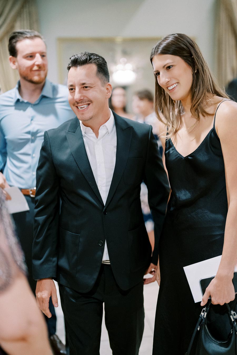 Group of people at a formal event, featuring a smiling man in a dark suit and white shirt standing beside a woman in a black dress, holding hands.