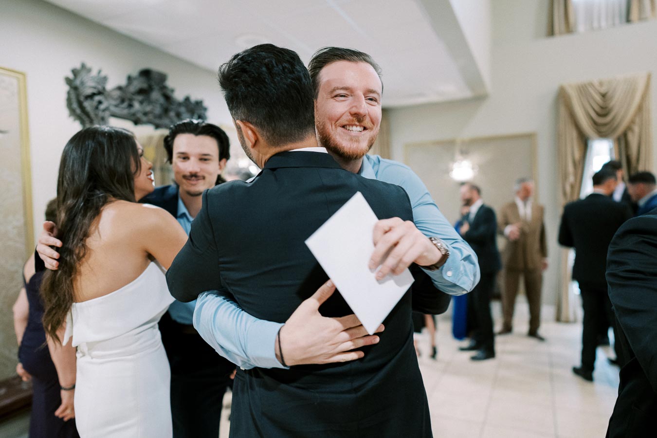 A group of people warmly hugging at a social event, with a man in a blue shirt smiling and holding an envelope, surrounded by elegantly dressed attendees.