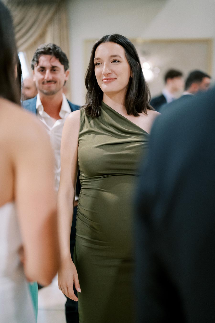 A woman in a stylish green dress attends a formal event, with people mingling in the background. She is smiling and engaged in conversation in a well-lit setting.