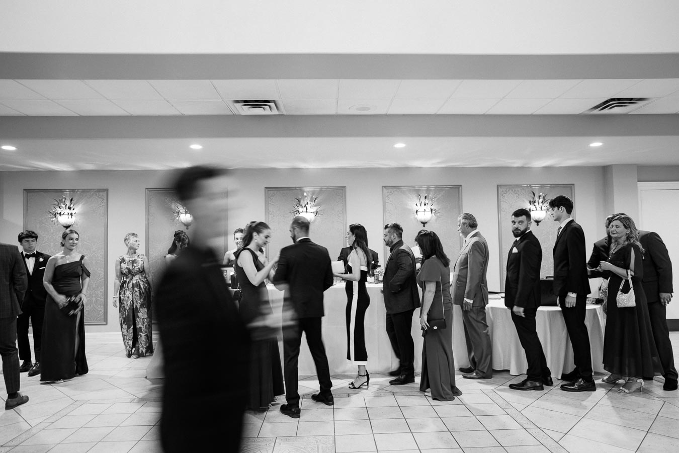 Black and white image of elegantly dressed people in formal attire lining up at a reception area during an event, with blurred motion capturing the dynamic atmosphere.