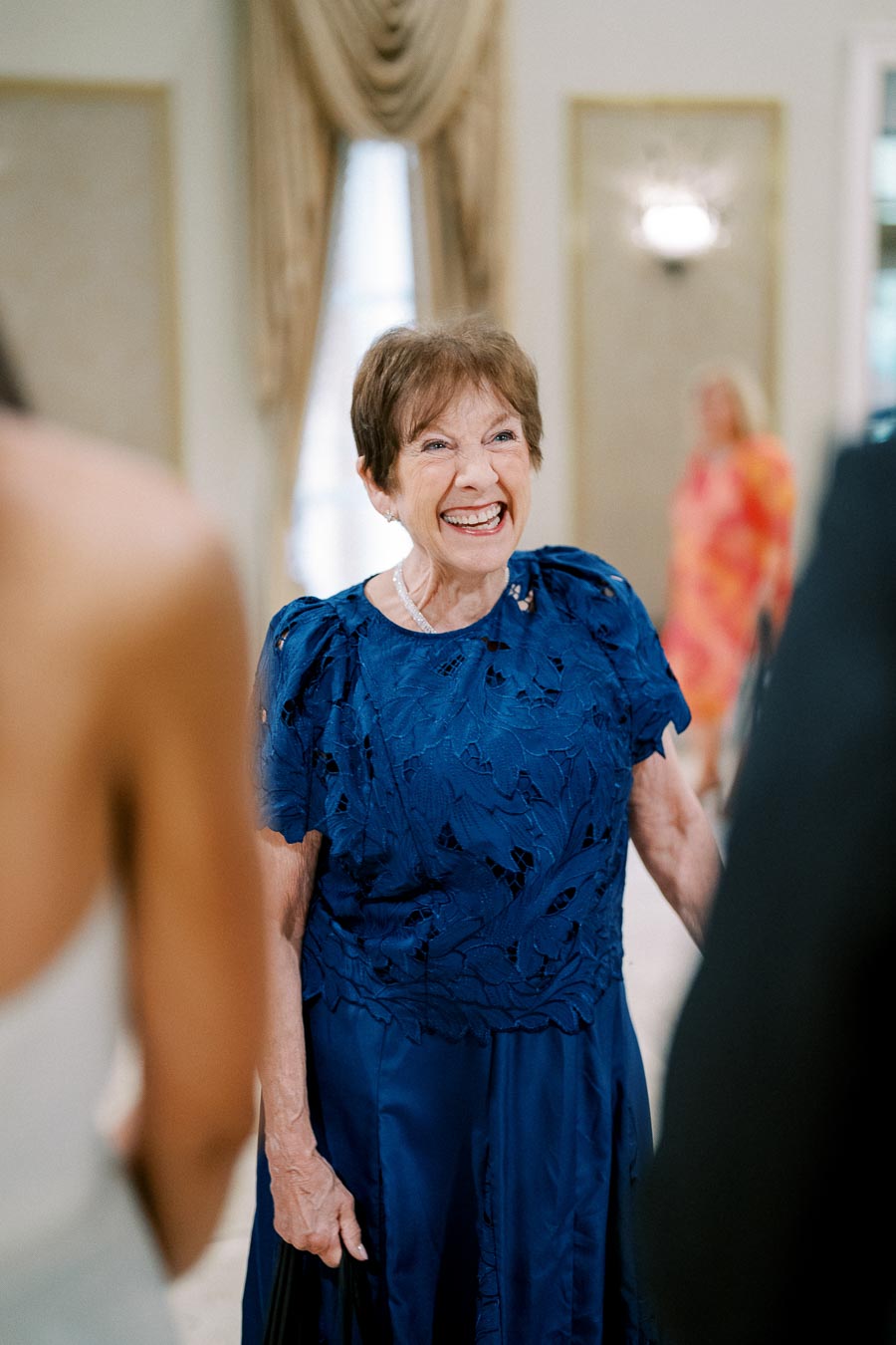 A joyful elderly woman in a blue dress smiling during a social gathering indoors, with blurred people and elegant decor in the background.