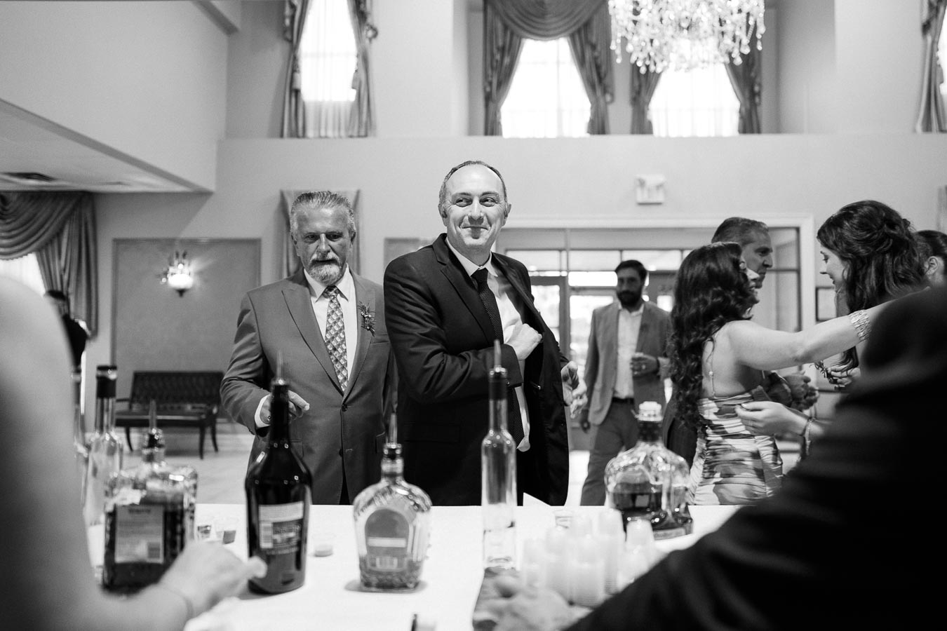 A group of elegantly dressed people socializing at an indoor event, with a variety of liquor bottles on a table in the foreground.