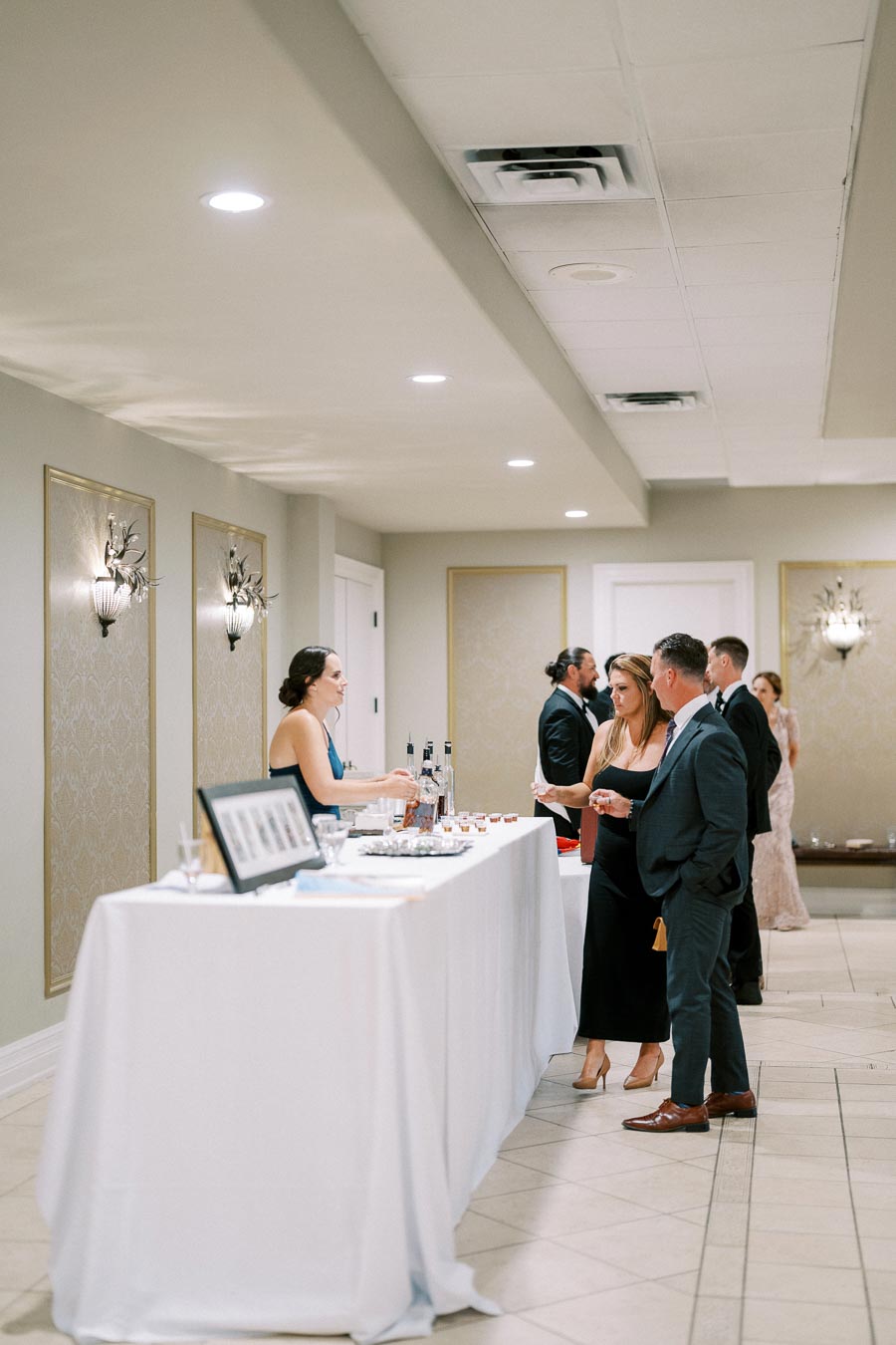 Elegant indoor event with guests interacting at a well-lit bar station, featuring white tablecloths and decorative wall sconces in a sophisticated venue setting.