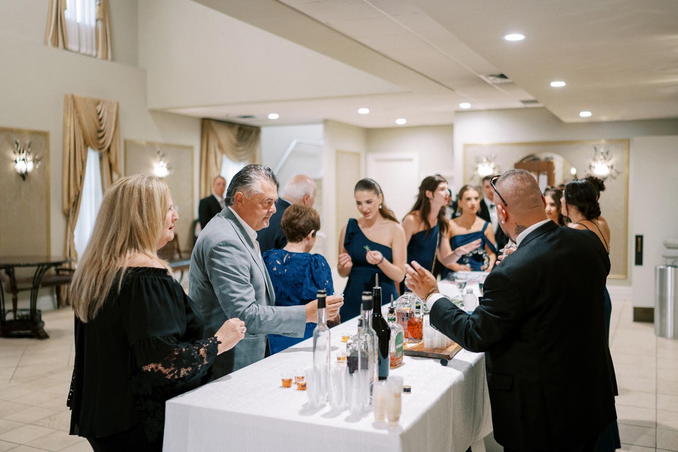 A group of elegantly dressed people socializing at a bar during a formal event, with bottles of liquor and glasses on a white tablecloth, in a well-lit reception hall.
