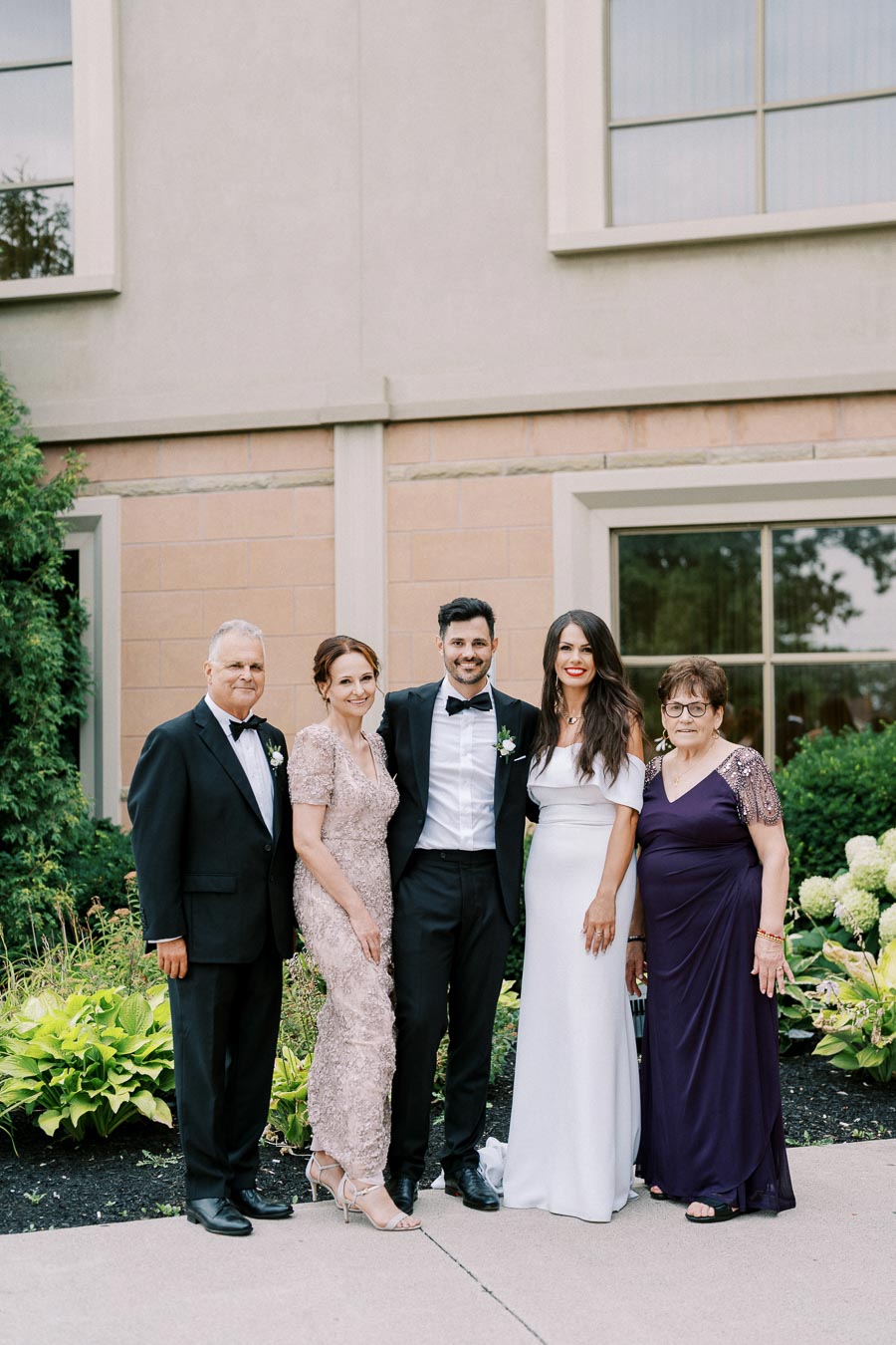 A family poses together in formal attire outside a building, featuring three women and two men dressed for a special occasion.
