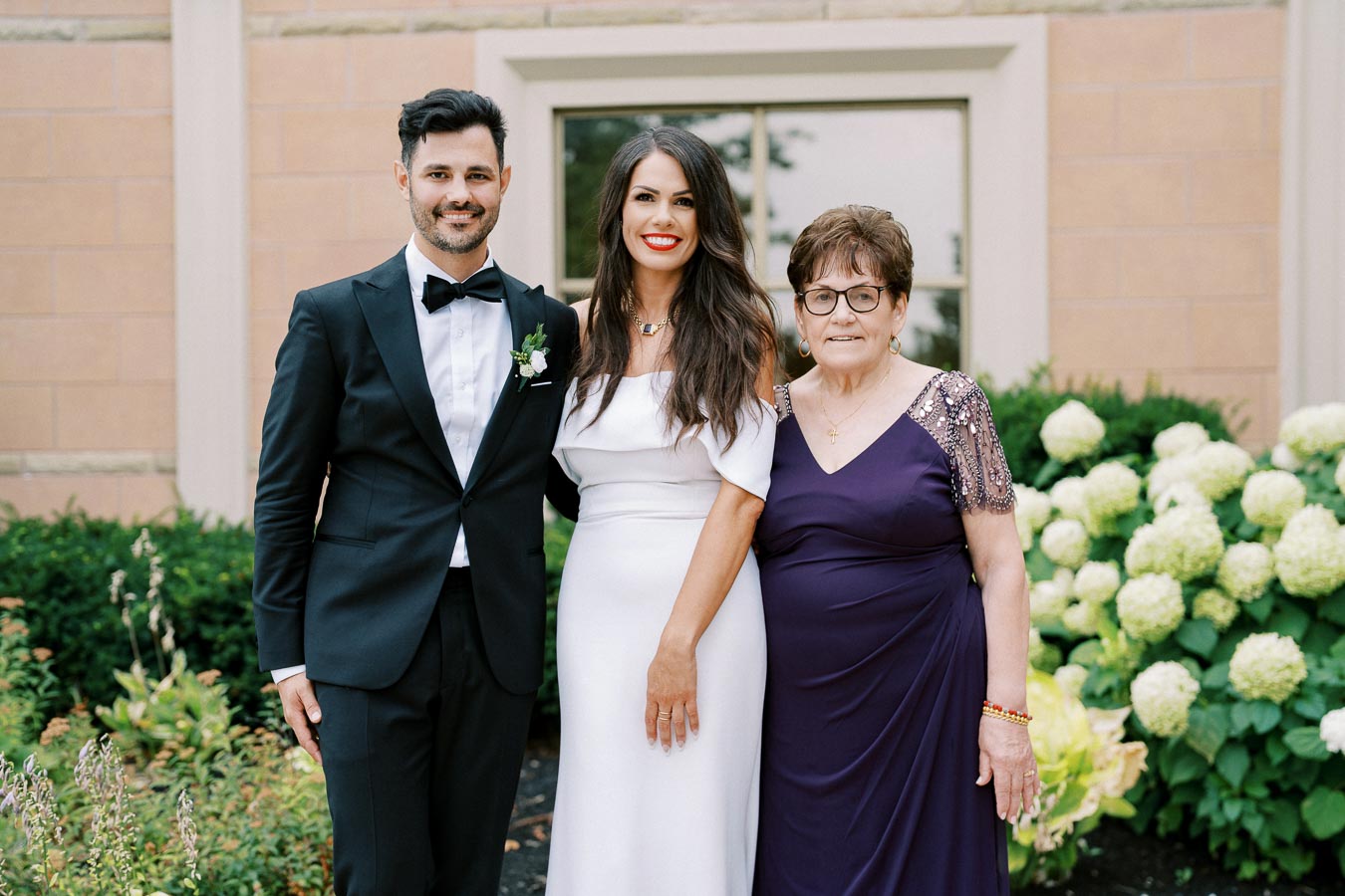 A groom in a black tuxedo, a bride in a white dress, and an older woman in a purple gown pose together outside near green bushes and a brick wall.