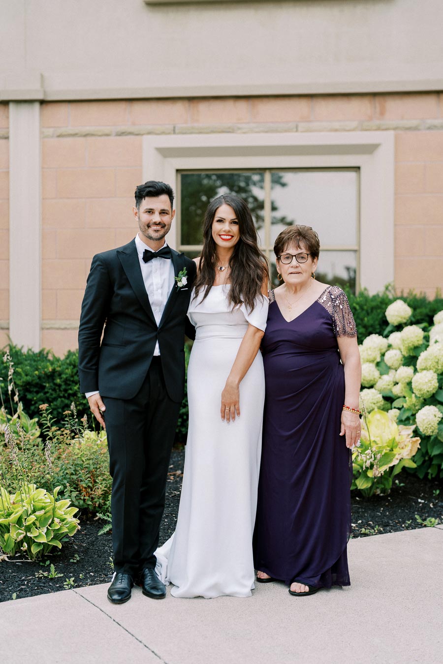 A groom in a black tuxedo and a bride in a white wedding dress posing outdoors with an older woman in a purple gown, surrounded by greenery and a building backdrop.