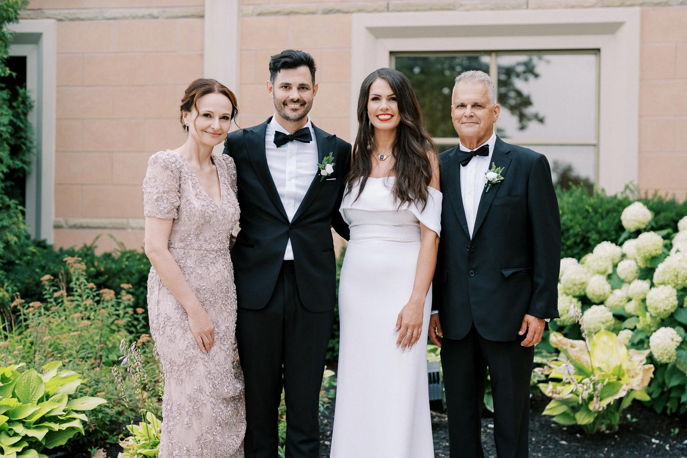 A group photo of four people dressed in formal attire, standing outdoors in front of a building with greenery and flowers. The men wear black tuxedos, and the women are in elegant dresses.
