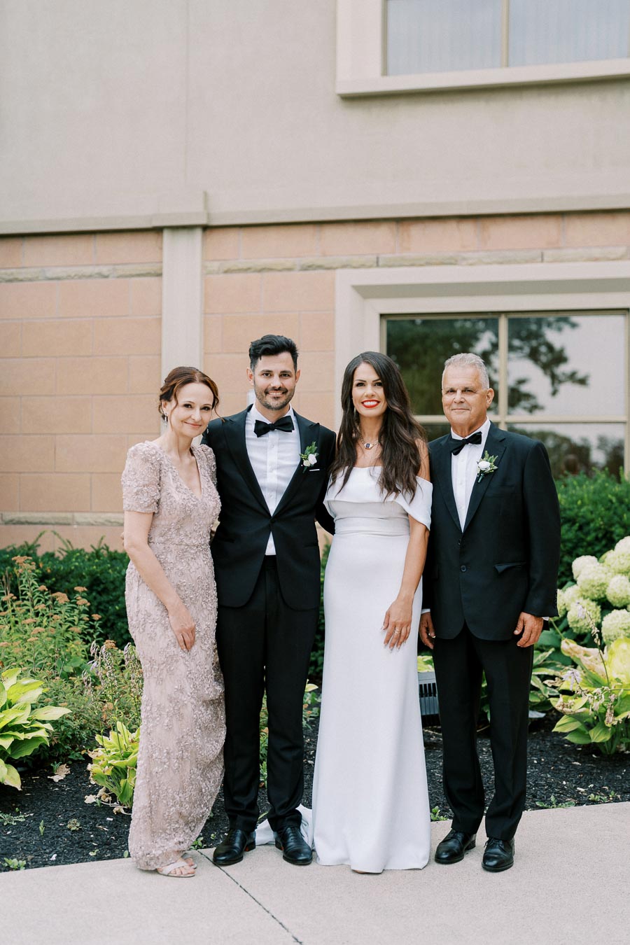 A bride and groom stand with their parents outside a building, dressed in formal attire. The bride wears a white dress, and the groom dons a black tuxedo. The group smiles amid landscaped greenery.