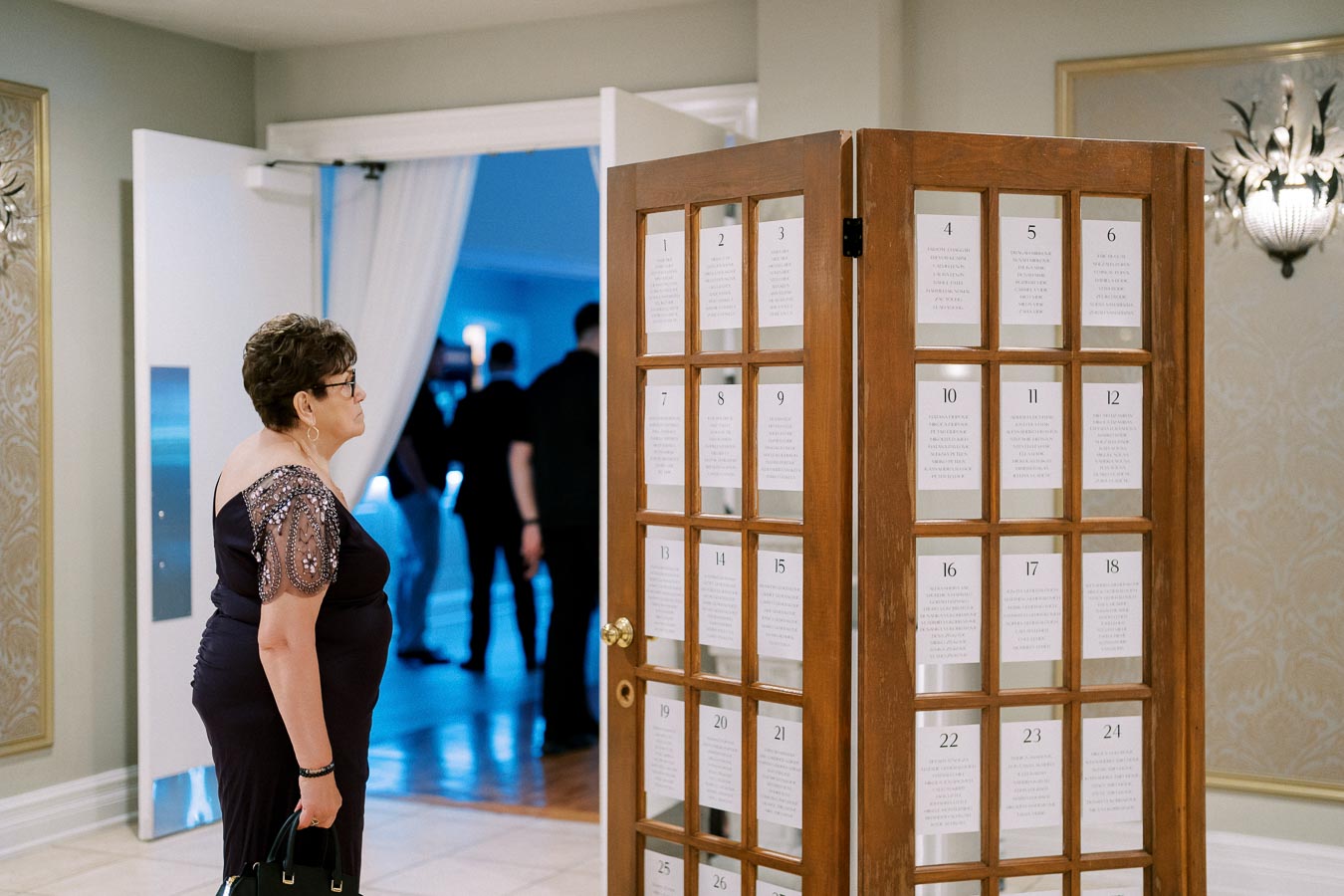 A woman in an elegant evening dress looking at a wooden door with numbered seating assignment cards, during a formal event in a venue with soft ambient lighting.