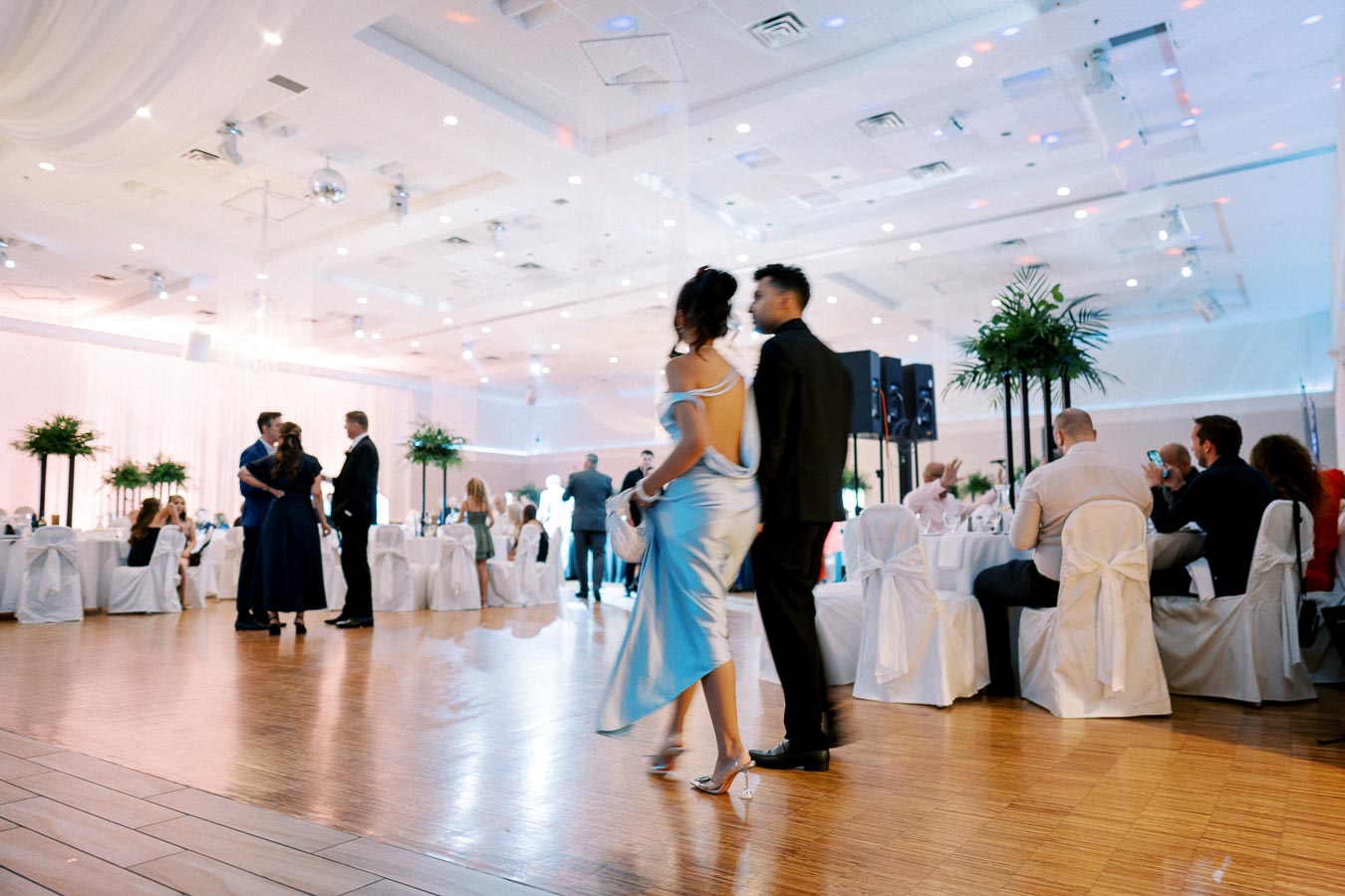 Elegant ballroom event with guests socializing; woman in blue dress and man in black suit walk across polished wooden dance floor surrounded by elegantly decorated tables with white drapes and green centerpieces.