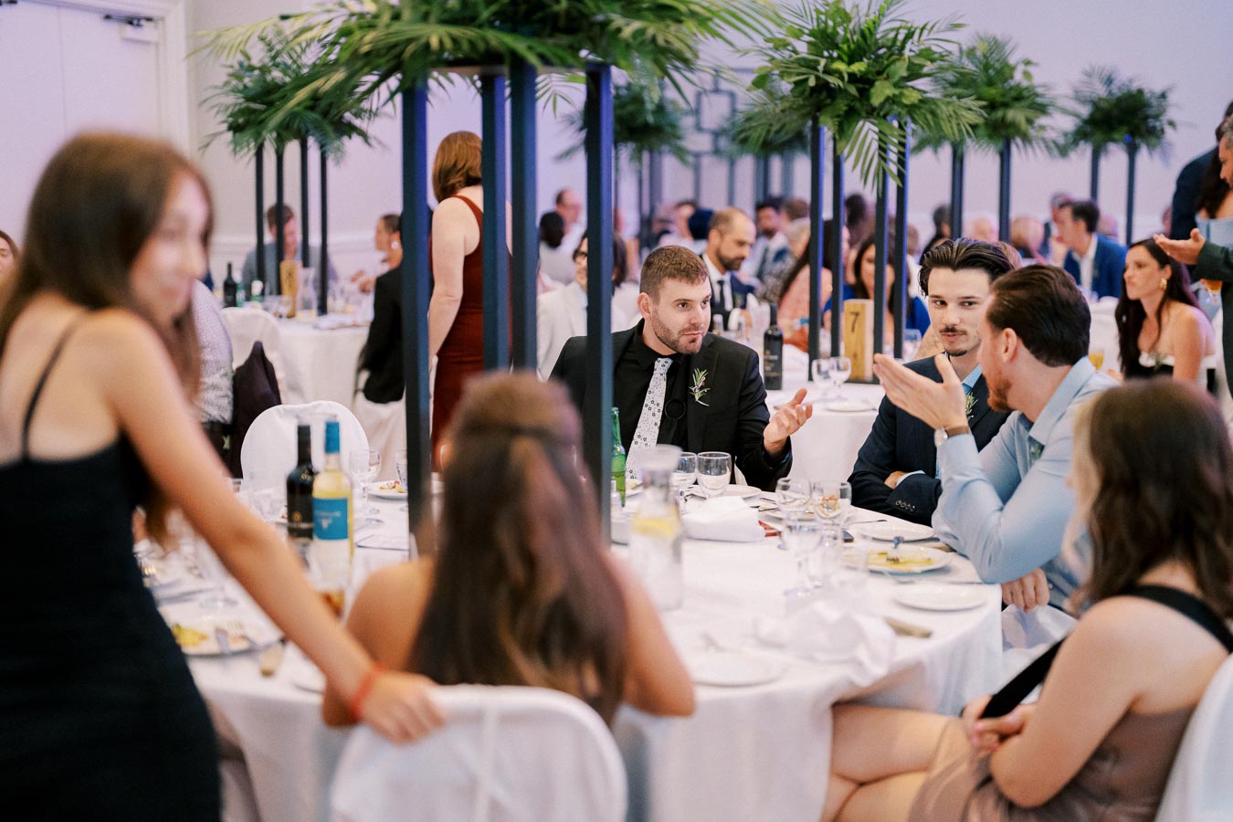 Group of people engaged in conversation at a formal event, seated around elegantly decorated tables with wine bottles and greenery centerpieces.