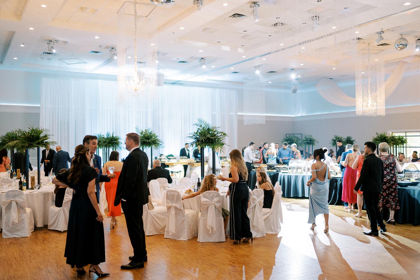 Elegant wedding reception in a beautifully decorated banquet hall, featuring guests mingling and dining, white draped chairs, round tables, and vibrant floral centerpieces under ambient lighting.