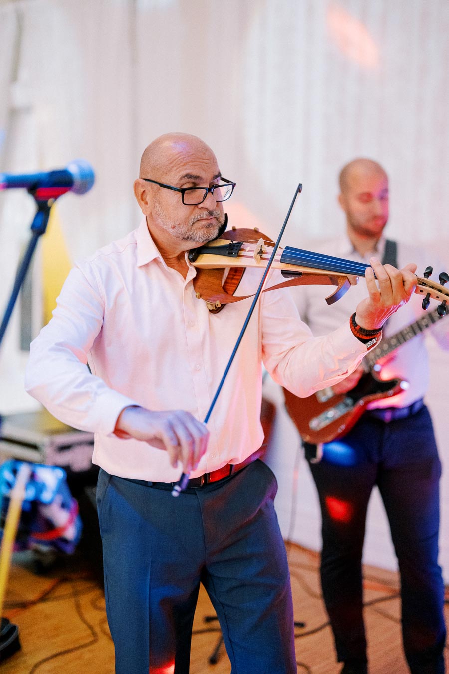 Man playing violin at an indoor event with guitarist in the background, featuring vibrant stage lighting and musical performance ambiance.