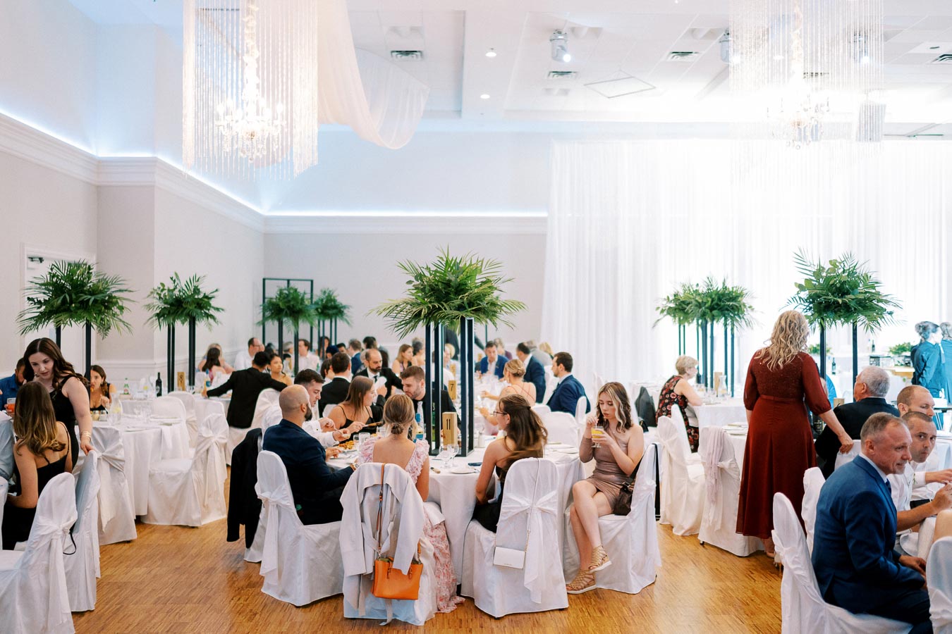 Elegant wedding reception with guests seated at round tables, adorned with white tablecloths and greenery centerpieces, in a spacious hall with chandeliers and ambient lighting.