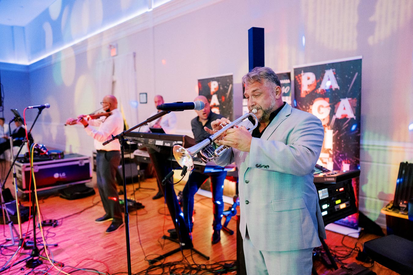 A live band performing at an indoor event, featuring a trumpeter in a light suit, a violinist, and a keyboardist. Colorful stage lights create a vibrant atmosphere, highlighting the musicians and their instruments.