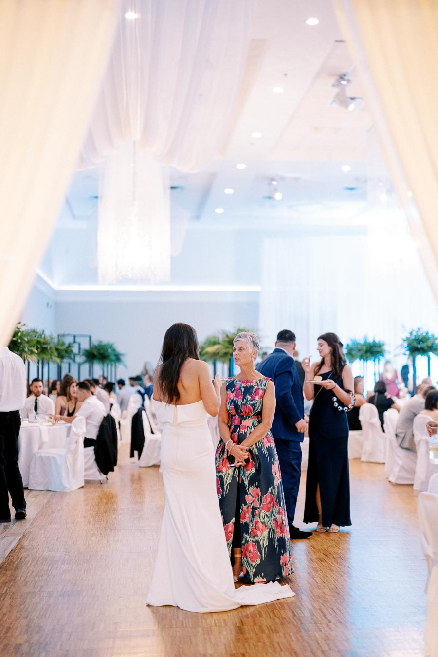 Elegant wedding reception with guests conversing in a beautifully decorated venue featuring white drapery, chandeliers, and floral arrangements.