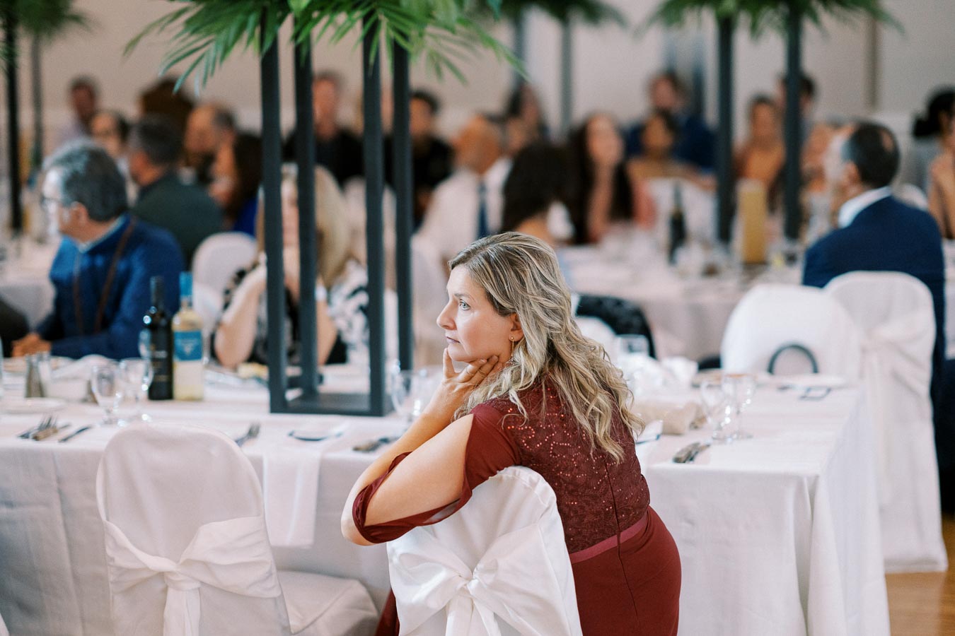 Elegant banquet scene with a woman in a red dress sitting at a table, overlooking a formal event with white tablecloths and decorative greenery.