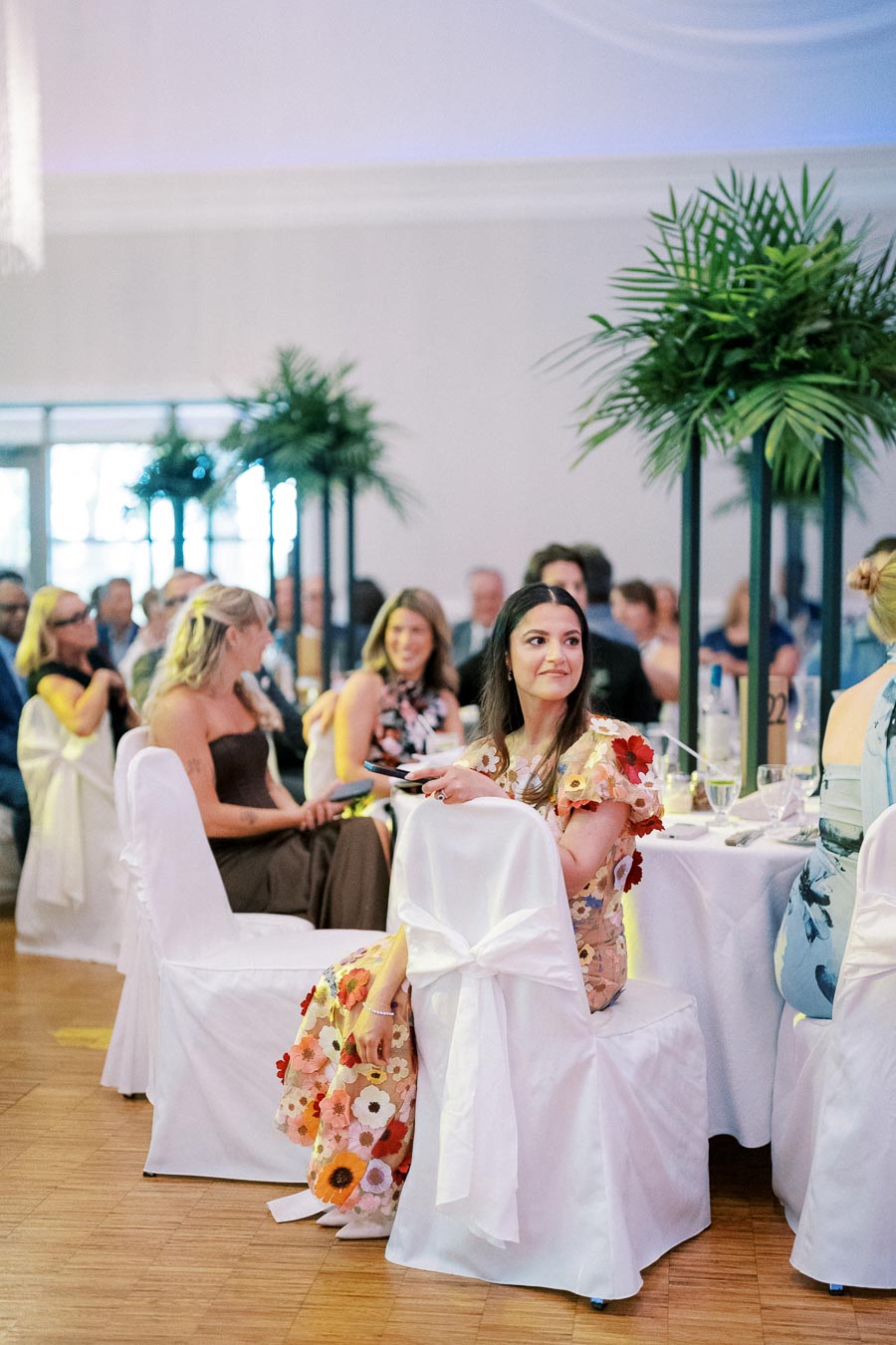Elegant wedding reception with guests seated around tables, featuring floral decorations, white chair covers, and tall green centerpieces in a beautifully lit venue.